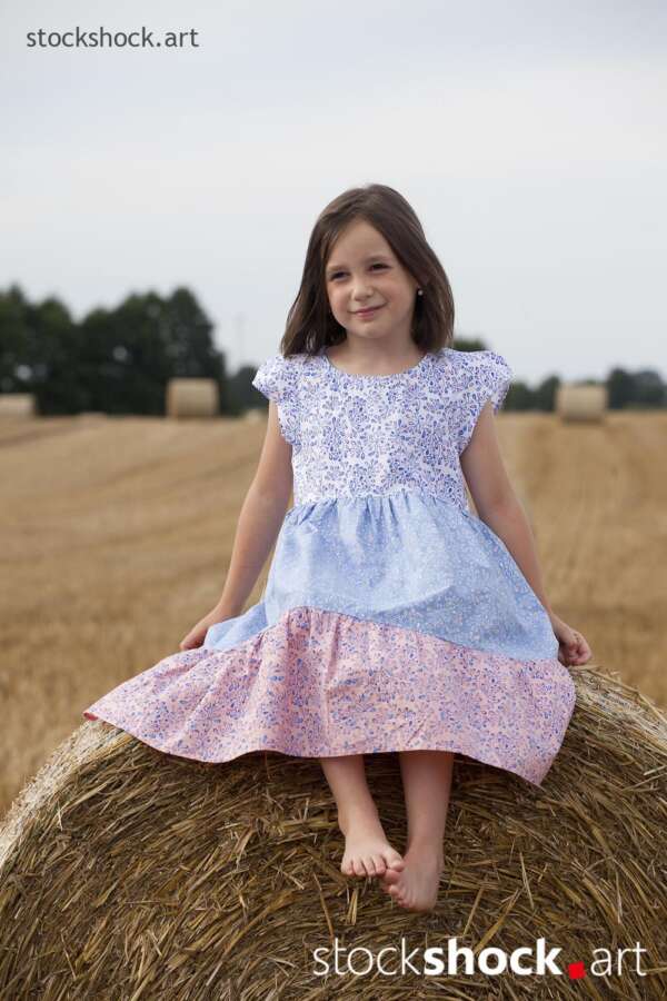 Girl sitting on a bale of straw in a field during harvest - stock image