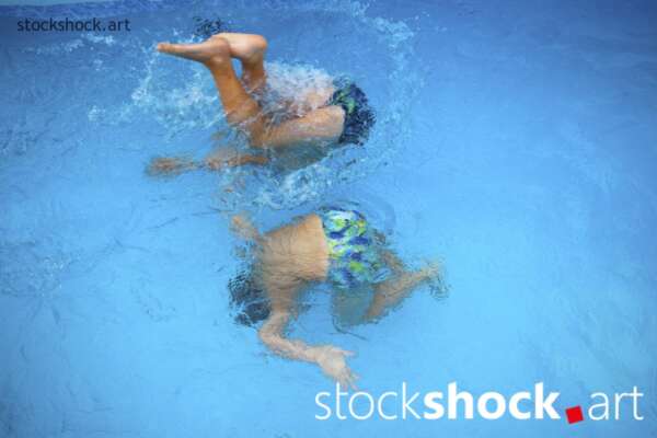 two brothers dive into the blue water of a pool, only his feet visible above the water