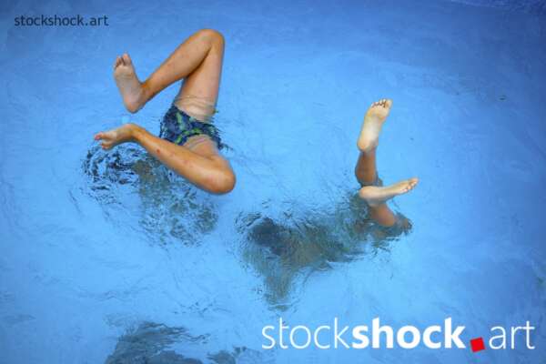 two brothers dive into the blue water of a pool, only his feet visible above the water
